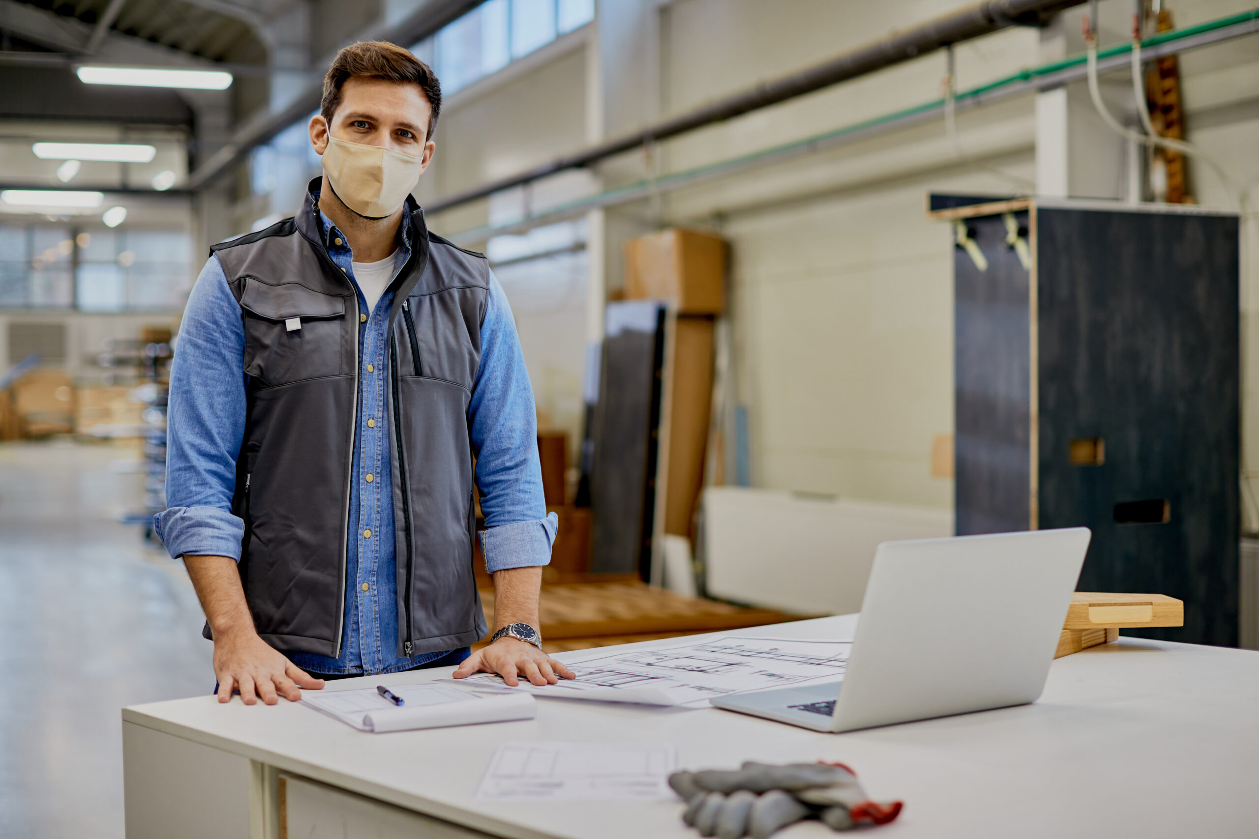 Male carpenter of woodworking factory wearing protective face mask while working at his desk and looking at camera.